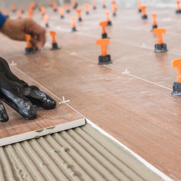 Worker installing wood-look ceramic floor tiles using spacers and gloves.