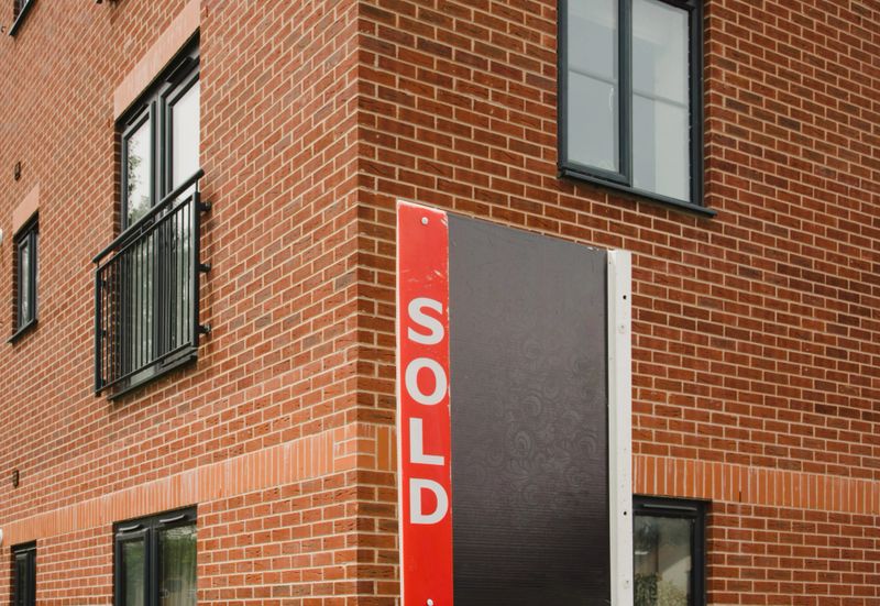 Estate agent sold sign in front of a red brick residential building of flats. Close view. Urban street. Housing problems in England. Property market in UK.