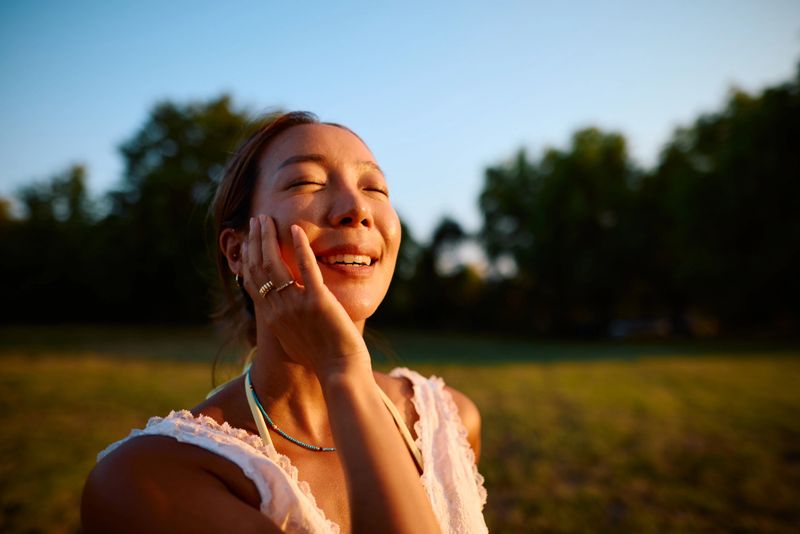 Young woman standing in a serene park at sunset, gently touching her face and smiling, surrounded by lush trees that enhance the tranquil atmosphere of golden hour