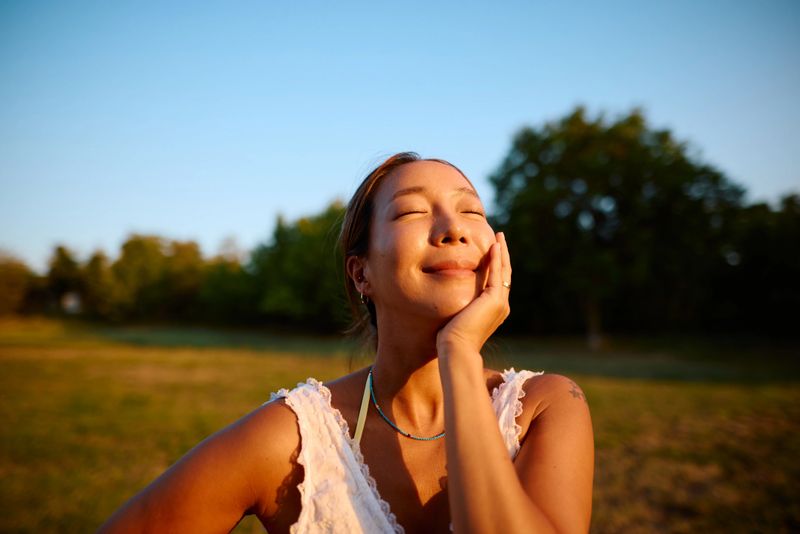 Serene woman with eyes closed, gently touching her face while basking in the warm glow of golden hour sunlight, enjoying the tranquility of a peaceful park setting