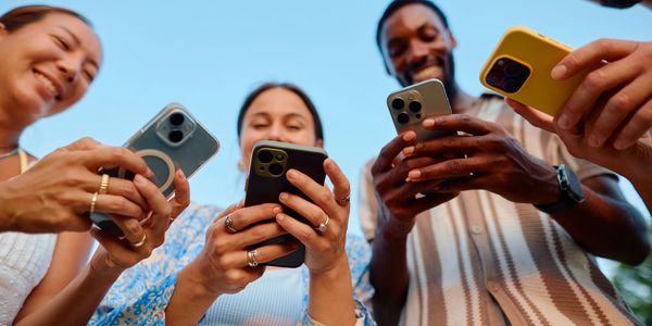 Four diverse friends smiling and using smartphones outdoors against a clear blue sky.