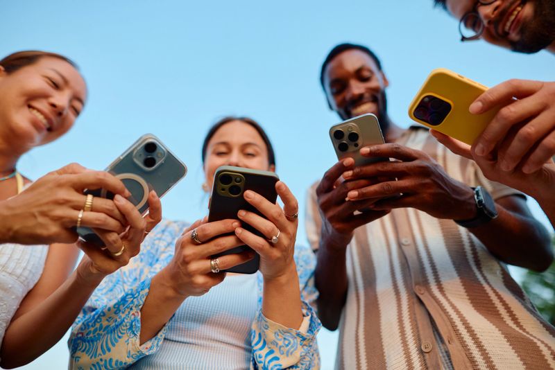 Four young adults smiling and enjoying each other's company while looking at their smartphones outdoors on a bright, sunny day, capturing moments of fun and connection