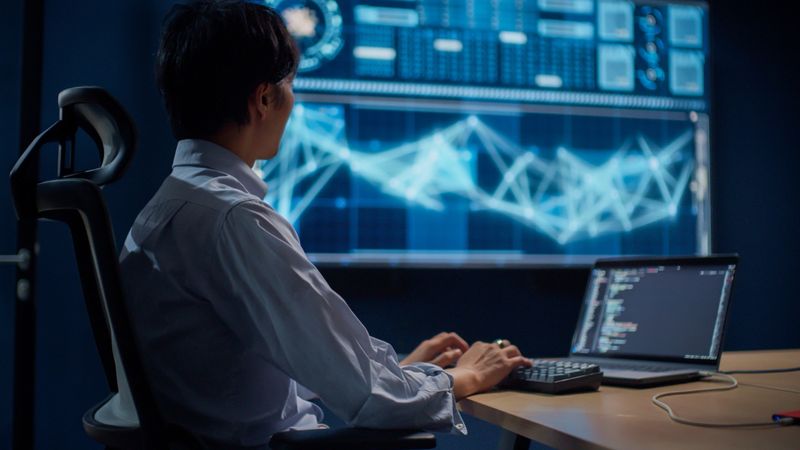 Artificial intelligence developer working in a modern dark-themed tech workspace. A male programmer sits in front of a large digital display showing data visualization and a laptop screen with lines of code. The image captures a realistic AI development environment, focusing on programming, machine learning, and teamwork in software engineering.