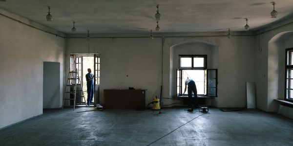 Two workers renovating a large empty room with windows and door open.