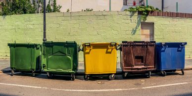 A row of colorful trash bins lined up against a green wall on a sunny street.