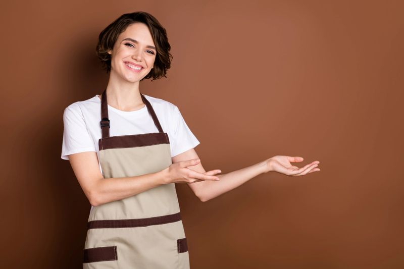 Portrait of attractive glad cheerful girl barista coffee shop demonstrating copy space welcome isolated over brown color background