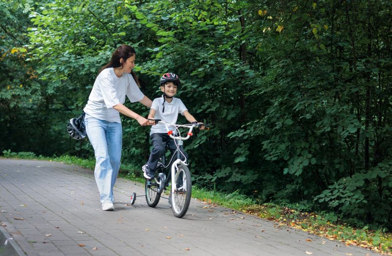 A caring mother helps her young son learn to ride a bicycle with training wheels in a green park. Concept of family support, maternal love, childhood development, parenting and active outdoor lifestyle.