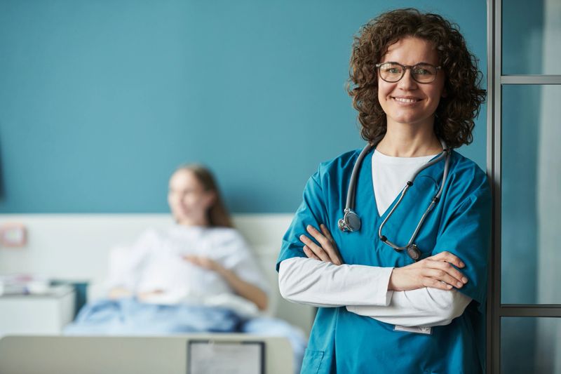 Portrait of middle aged Caucasian woman doctor with curly hair standing with arms crossed and smiling, stethoscope around neck, patient lying in hospital bed in background