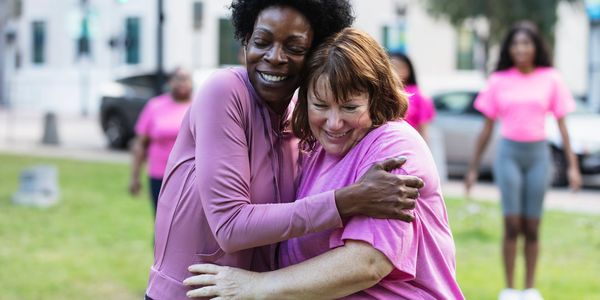 Two women happily hugging outdoors with others in pink shirts nearby.