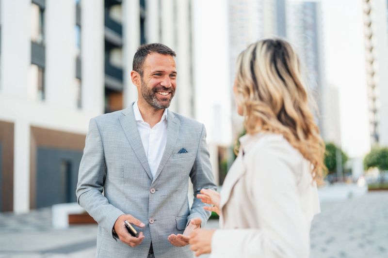 Cheerful business persons discussing new project and sharing ideas while walking in front of their office building. Real estate agent and a client are talking in front of a tall modern building in the city.