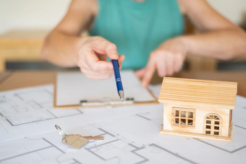 Real estate agent pointing with a pen at the signature line on a home purchase agreement, surrounded by house keys and a small wooden model house resting on blueprints