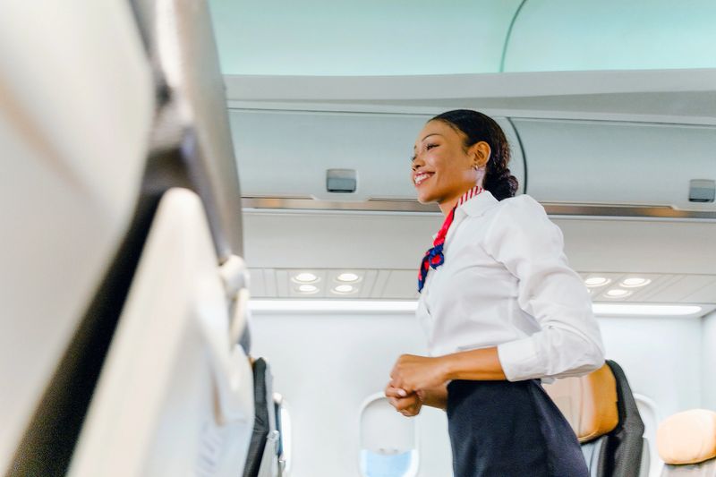 Smiling flight attendant in uniform standing in an airplane cabin aisle, offering service to passengers during boarding.