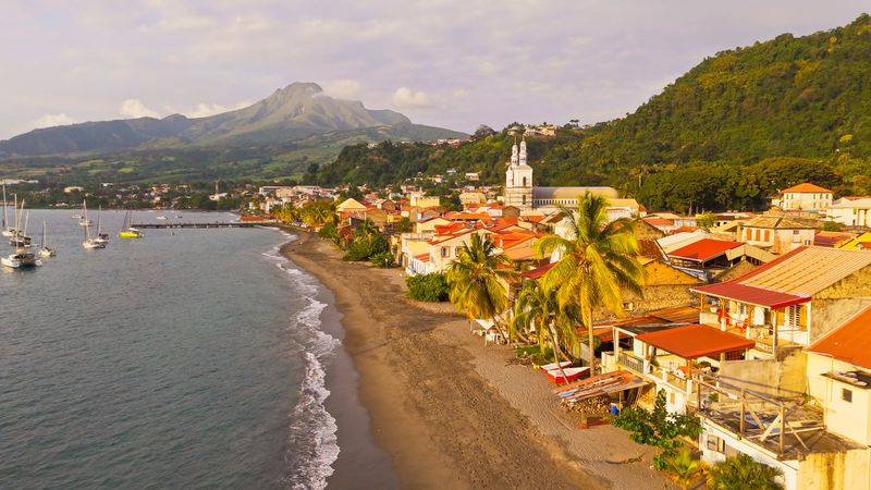 A charming coastal town in Dominica is featuring vibrant buildings along a sandy beach. Sailboats are anchored nearby on the calm sea, while lush green mountains are rising in the background under a partly cloudy sky.