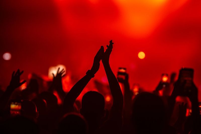 Silhouette of fans clapping and filming with smartphones at a live music concert under red stage lights