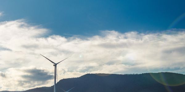 Wind turbines stand tall against a blue sky with clouds over a mountain.