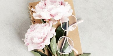 Pink peonies, glasses, and a notebook on a gray surface.