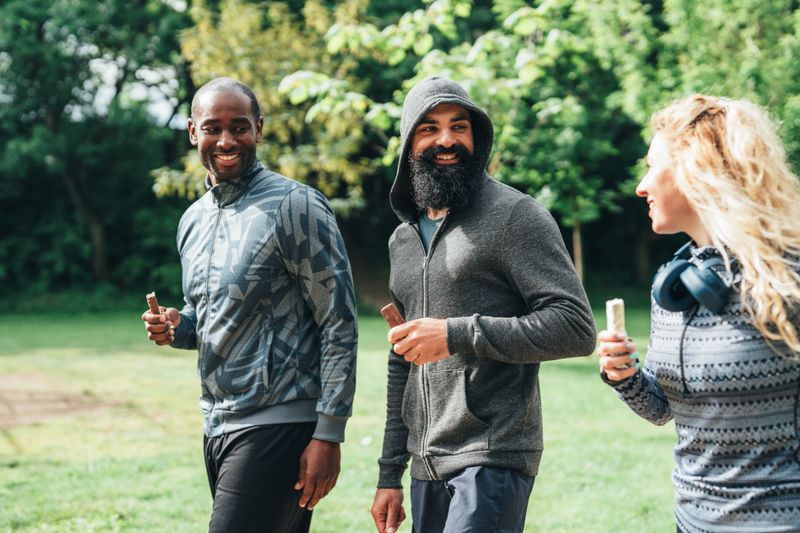 Three fit friends walking together in a park after exercising, smiling and enjoying protein healthy bars. The group, dressed in athletic wear, appears relaxed and cheerful, highlighting a healthy and active lifestyle. The scene captures post-workout camaraderie and the importance of nutrition and friendship in fitness routines.