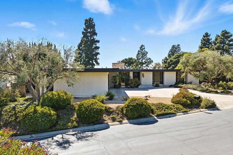 Mid-century modern house with lush landscaping under a bright blue sky, featuring trees and shrubs in a suburban setting.