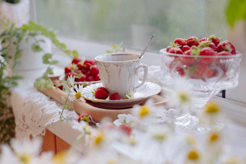 A beautiful kitchen setting features a cup of tea placed on a wooden tray with fresh strawberries. Delicate flowers and a crystal bowl of berries add charm to the bright ambiance.