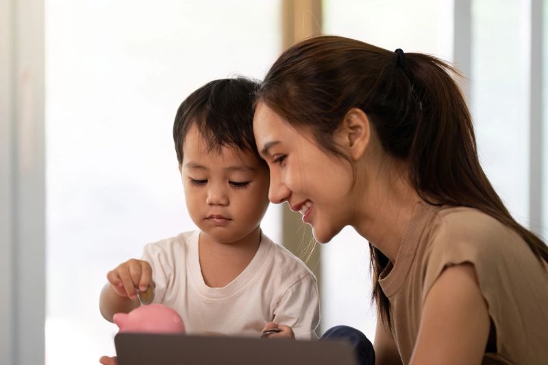 A mother engages her young son in financial literacy by using a piggy bank, promoting saving habits in a cozy home environment.