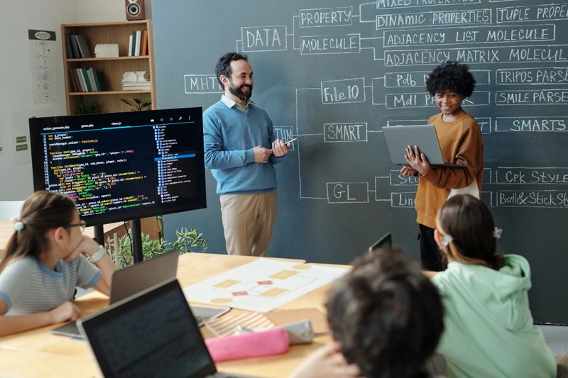 Teacher engaging diverse group of students with programming on whiteboard and monitor, all immersed in learning. Classroom environment includes computers and educational materials