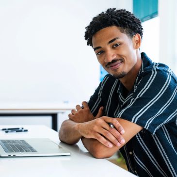 Homem sorridente sentado em mesa com laptop.