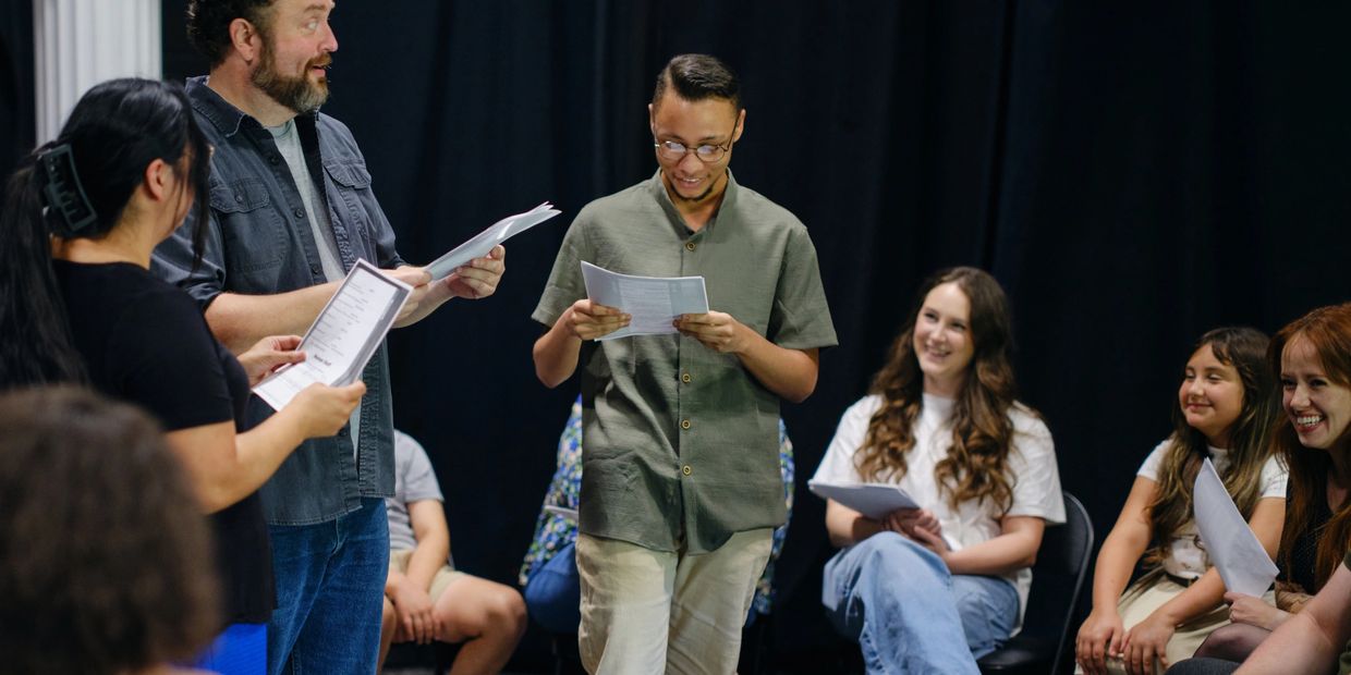 Three actors stand on stage holding scripts with other actors seated around them.