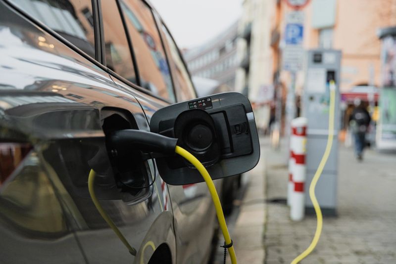 A modern electric vehicle is parked and plugged into a public charging station on a street in Berlin, Germany. The scene captures the growing trend of sustainable urban transportation, showcasing a clean-energy solution in a contemporary European city environment. Yellow charging cables and clear reflections on the vehicle emphasize the sleek design and urban integration of EV technology.