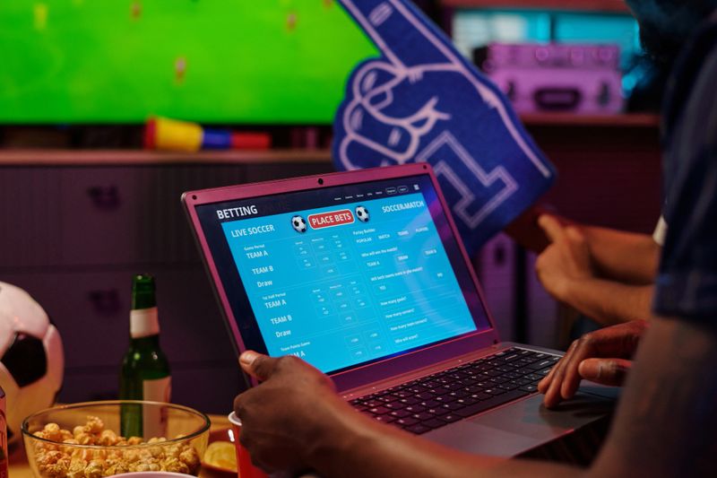 Black man using laptop for online sports betting during soccer match, hands visible interacting with screen showing live soccer odds, snacks and drink on table in foreground