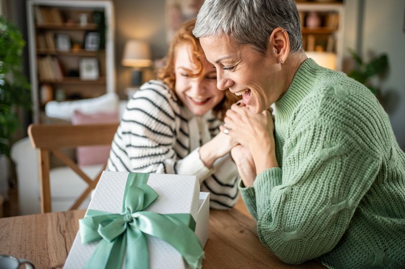 Two mature women are sitting at a table, sharing a tender moment as they admire a beautifully wrapped gift, filled with love and anticipation