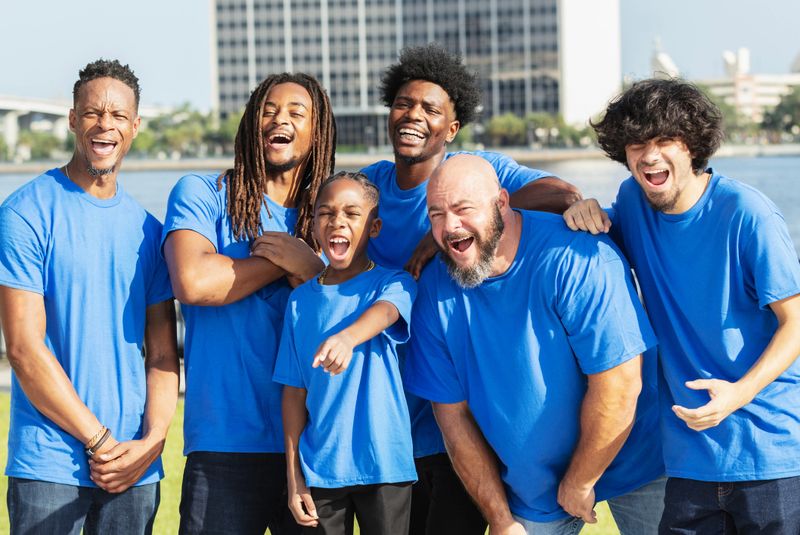 A multiracial group of men and a boy wearing blue t-shirts, a team standing together on a city waterfront. They are all looking in the same direction, laughing, amazed and excited about what they are watching. They are family and friends, fathers and sons.