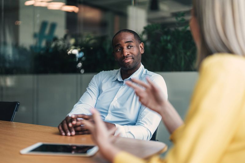 Confident male advisor having a professional discussion with a client in a modern office environment. The setting suggests a one-on-one consultation, with documents and a digital device on the table, highlighting themes of business strategy, financial planning, or client relationship management.