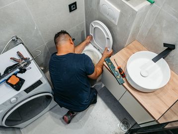 A man repairs a toilet in a modern bathroom with tools nearby.