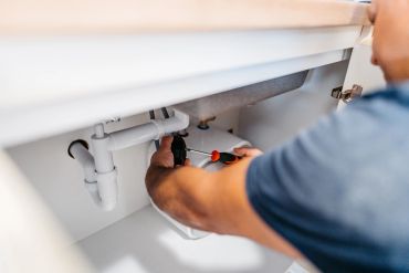 A person fixing plumbing under a kitchen sink with a screwdriver.