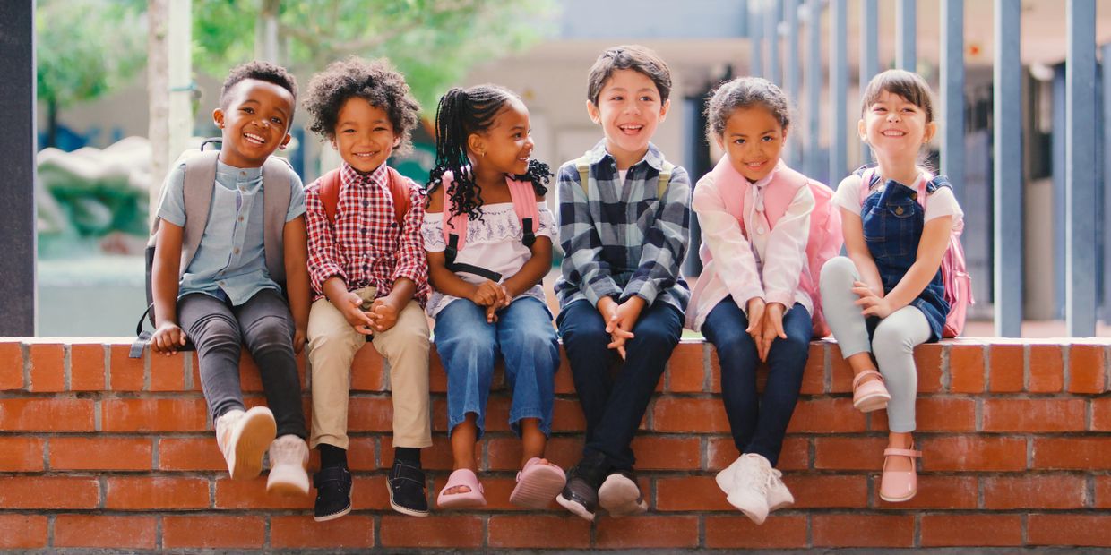 Six diverse children with backpacks sitting happily on a brick wall outdoors.