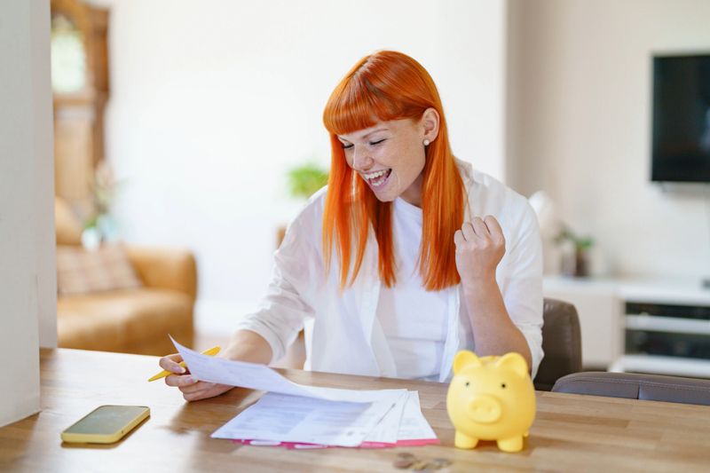 A joyful woman with vibrant orange hair raises her fist in excitement while looking at financial documents at her kitchen table, featuring a piggy bank.