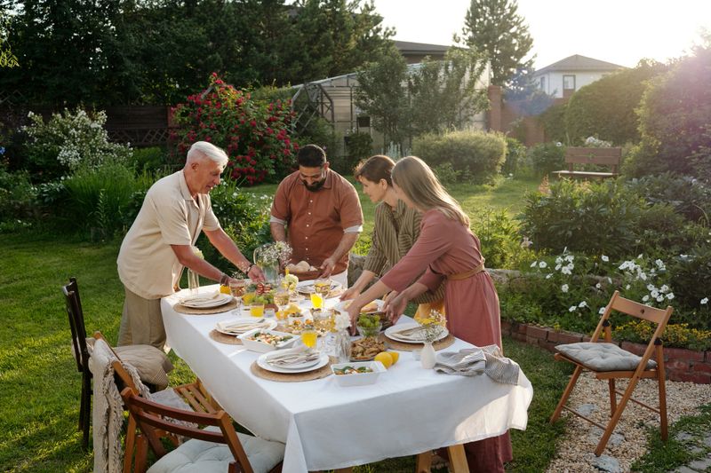 Caucasian senior man, Middle Eastern man, Caucasian middle aged woman, Caucasian young adult woman arranging food and drinks on outdoor dining table in garden during daytime gathering
