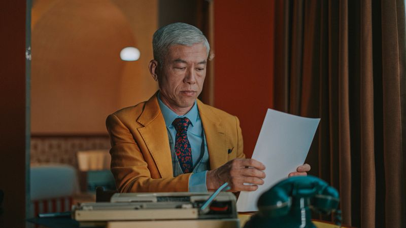 senior chinese man in a suit reviews a typed page from a vintage typewriter at a desk with a classic rotary phone beside him, lit by soft window light in a retro office setting. Location: indoor.