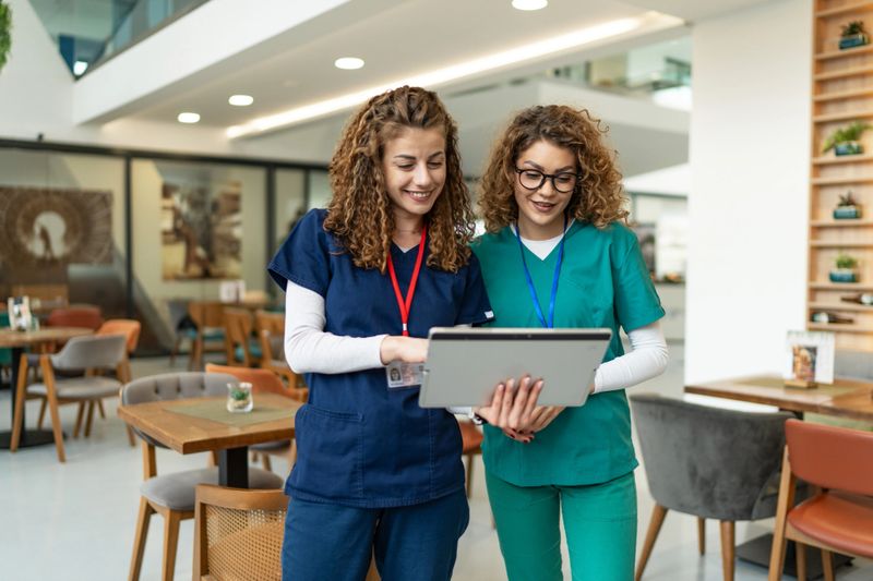 Two smiling female nurses in scrubs are standing in the hospital cafeteria, looking at a digital tablet and discussing patient information during their break