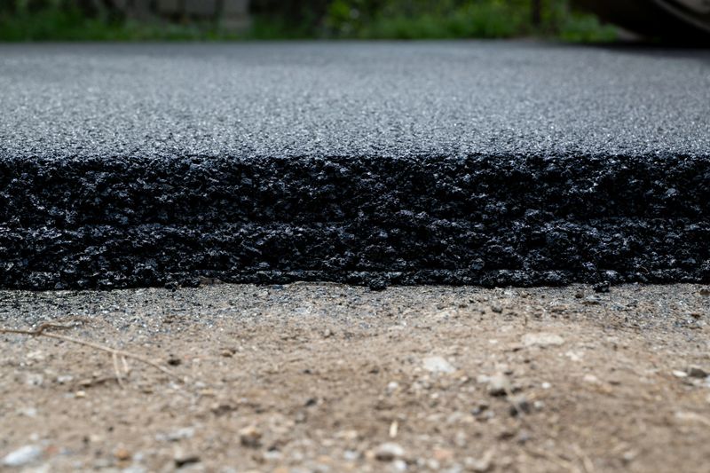 A detailed close-up shows the edge of a freshly laid asphalt layer on a road surface, revealing the texture of compacted blacktop against the rough ground beneath. The image emphasizes infrastructure development, construction quality, and the durability of modern roadwork.