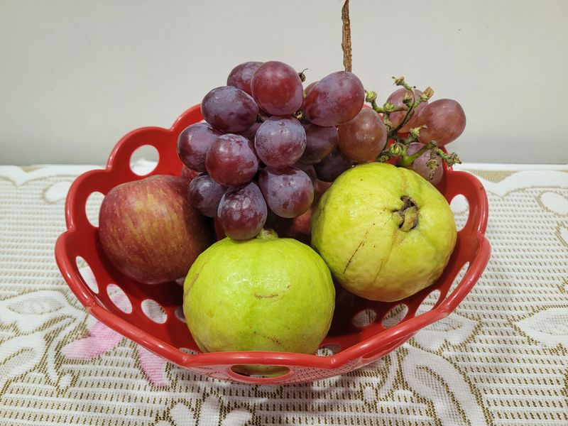 Vibrant display of fresh apples, purple grapes, and quinces arranged in a red fruit bowl. The colorful fruits are neatly placed on a tablecloth, offering a refreshing and inviting look that highlights the natural beauty and variety of seasonal fruits.