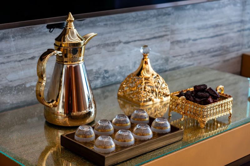 In the photo, a golden Arabic coffee pot stands on a table with dates and cups. A traditional scene typical for rich Middle Eastern countries like Qatar, UAE, and Saudi Arabia.