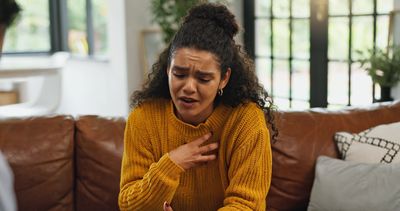 A woman in a mustard sweater clutching her chest in discomfort on a couch.