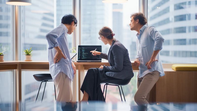 Three software engineers, including two men and one woman, are gathered around a laptop discussing programming code in a modern, high-rise office with large windows. The team appears to be reviewing or debugging software together, highlighting teamwork, collaboration, and the creative process in a tech startup or IT company. The cityscape outside the windows suggests a professional urban environment. This image conveys concepts of software development, agile workflow, modern business, innovation, and corporate teamwork.