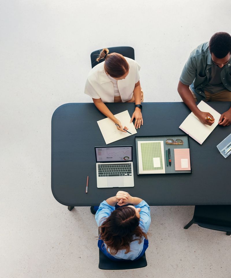 High angle shot of a group of professionals engaged in a business meeting with notepads and a laptop on the table, emphasizing teamwork and productivity in a corporate environment.