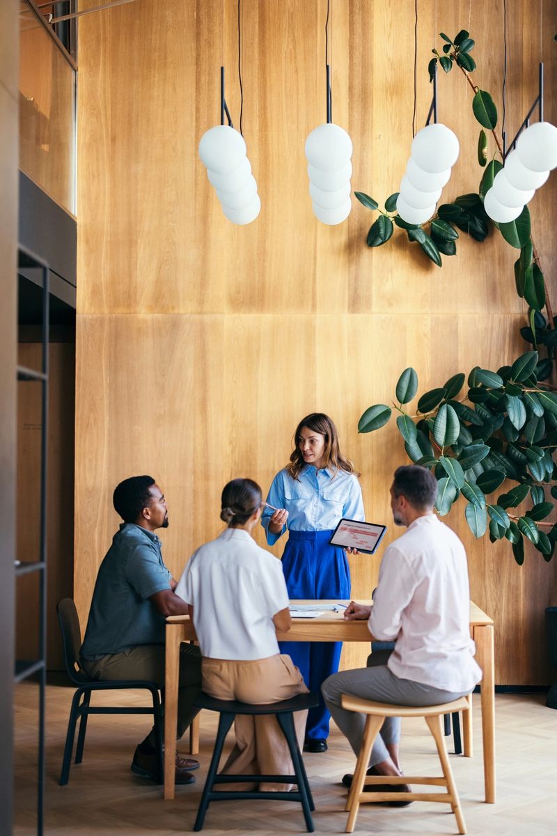 A group of coworkers collaborates at a table in a stylish office environment. The space features a wooden interior, modern lighting, and greenery, fostering creativity, teamwork, and a productive atmosphere.