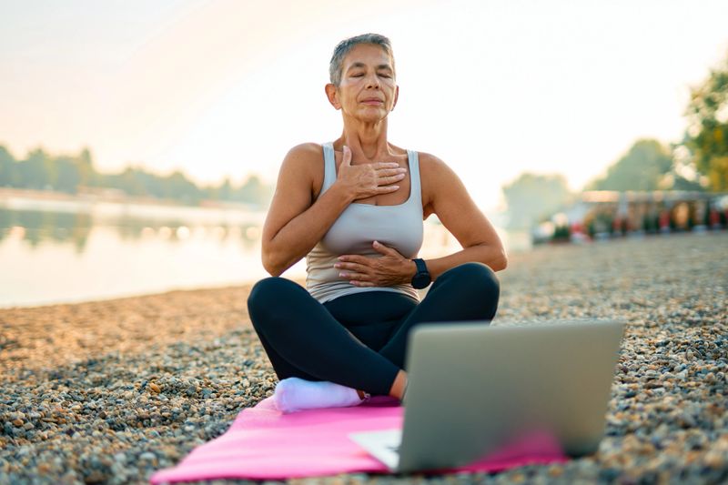 Mature woman is sitting on a yoga mat on a pebble beach by the river at sunrise, practicing breathing exercises with the help of an online course on her laptop