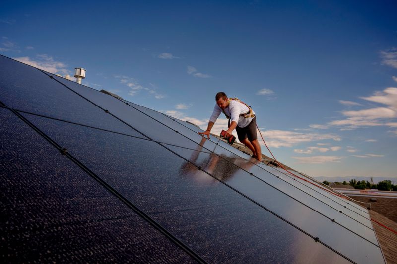 A residential solar energy system is being installed by a technician wearing safety gear and using a cordless drill. The scene takes place on a rooftop in a quiet suburban neighborhood, with blue skies and distant views of rooftops and mountains.