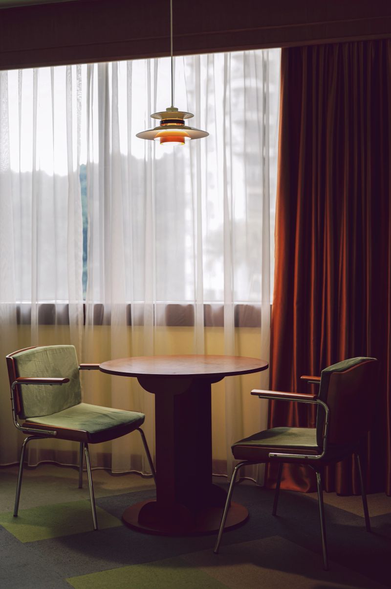 Minimalist interior design with two retro chairs and a round wooden table placed near a large curtained window, with soft natural light and a hanging pendant lamp creating a calm and stylish atmosphere.