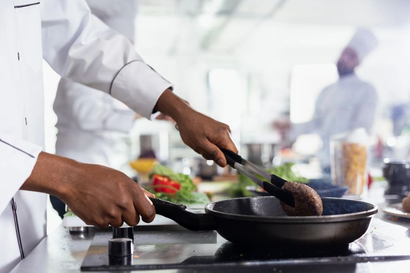 African american chef flipping the pork fillet in the pan using tongs, preparing the raw meat on the restaurant kitchen stove. Young male expert cooking steak in oil, food industry. Close up.